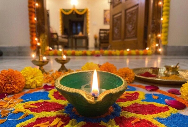 May include: A green clay oil lamp with a gold-painted rim and a lit flame, resting on a colorful circular floor pattern made of colored powder. The scene includes scattered orange and yellow marigold flowers and small brass bowls nearby.