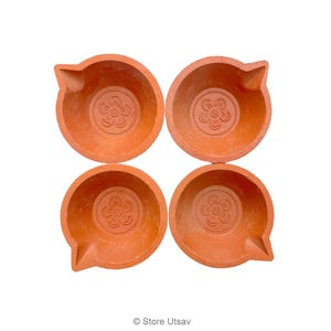 May include: Four terracotta oil lamps arranged in a square. Each lamp has a round bowl shape with a spout and a floral design in the center. The lamps are a warm orange-brown color, set against a white background.