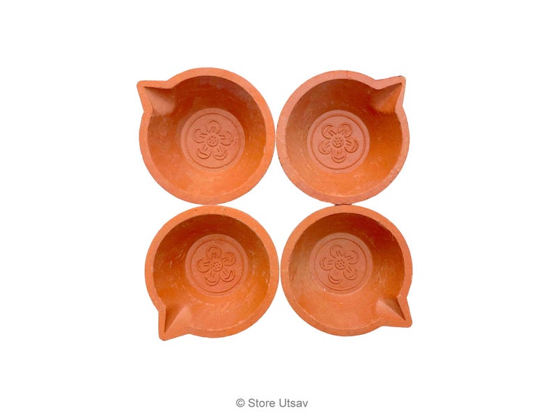 May include: Four terracotta oil lamps arranged in a square. Each lamp has a round bowl shape with a spout and a floral design in the center. The lamps are a warm orange-brown color, set against a white background.