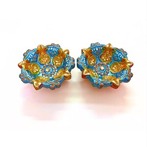 May include: Two small, round, hand-painted clay oil lamps featuring a vibrant blue base color with intricate, raised floral and paisley patterns highlighted in metallic gold and green paint. The edges of the bowls have a scalloped, petal-like shape.