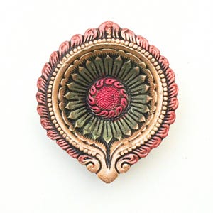 May include: A decorative oil lamp, or diya, featuring a circular shape with a pointed tip. The design includes a central textured red circle surrounded by green petals, encased in a metallic gold border with a scalloped, reddish-pink outer rim.