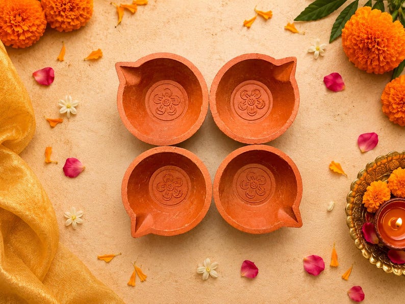May include: Four terracotta oil lamps with a floral design are arranged on a beige surface, surrounded by orange marigold flowers, petals, and a golden cloth. A small candle is lit in a decorative dish.