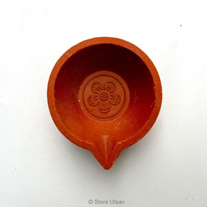 May include: A terracotta-colored, round oil lamp with a central floral design. The lamp has a small spout and a shallow bowl. The design is embossed into the clay. The lamp is likely used for religious or decorative purposes.