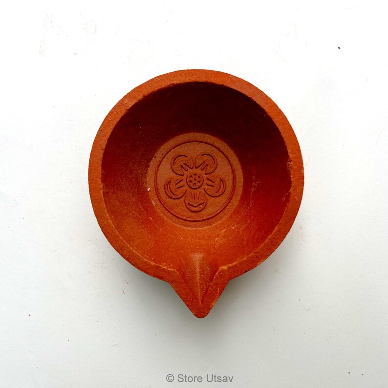 May include: A terracotta-colored, round oil lamp with a central floral design. The lamp has a small spout and a shallow bowl. The design is embossed into the clay. The lamp is likely used for religious or decorative purposes.