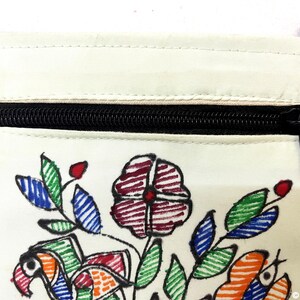 May include: A cream-colored fabric pouch featuring a hand-drawn floral and bird design in red, green, blue, and orange ink. The pouch has a black zippered closure at the top and is bordered by blue fabric trim along the sides and top edges.