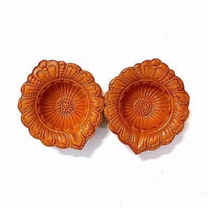 May include: Two terracotta oil lamps, known as diyas, featuring a circular shape with a scalloped petal-like edge and a central textured floral pattern. The clay material has a warm, earthy orange-brown tone with a matte finish.