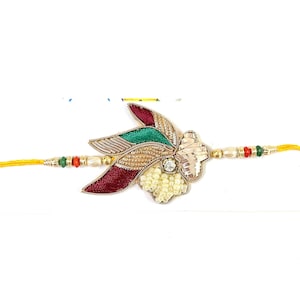 May include: A decorative bracelet featuring a central floral-inspired ornament with maroon, green, and gold metallic thread embroidery, accented by small white beads and a clear crystal, attached to a yellow cord with small red, green, and gold beads.