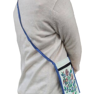 Woman wearing a small Madhubani painted sling bag. Its a light blue crossbody bag with a floral design. The bag has a mint blue background with a black zipper and a blue strap. The floral design includes pink, orange, and green elements.