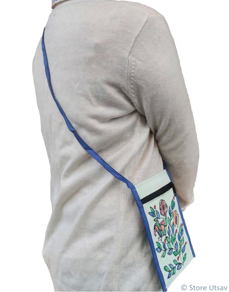Woman wearing a small Madhubani painted sling bag. Its a light blue crossbody bag with a floral design. The bag has a mint blue background with a black zipper and a blue strap. The floral design includes pink, orange, and green elements.
