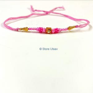 May include: A pink bracelet with a central square gold-colored bead with a small clear gem. The bracelet features pink beads and gold-colored accents. The text "Store Utsav" is visible.