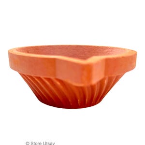May include: A terracotta-colored diya, a traditional Indian oil lamp, is shown. The lamp has a wide, open bowl shape with a pointed spout for the wick. The exterior features a ribbed design, and the overall form is symmetrical and simple.