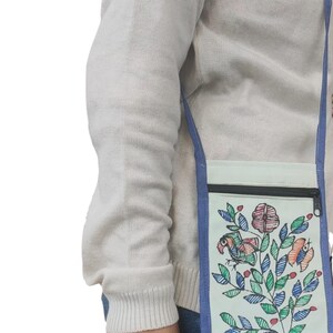 May include: A light-colored rectangular crossbody bag featuring a black zippered top closure and a blue fabric strap. The front of the bag displays a colorful hand-drawn illustration of a bird perched among leafy branches and flowers in shades of red, blue, and green.