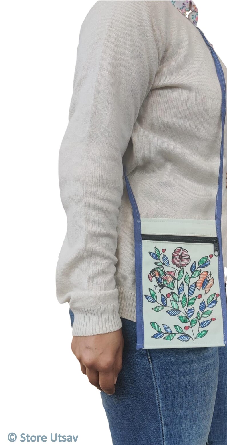 May include: A light-colored rectangular crossbody bag featuring a black zippered top closure and a blue fabric strap. The front of the bag displays a colorful hand-drawn illustration of a bird perched among leafy branches and flowers in shades of red, blue, and green.