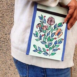 May include: A rectangular crossbody bag with a light green base, blue trim, and a black zipper closure. The front features a hand-drawn illustration of a bird, a flower, and a butterfly surrounded by green leaves and small red berries.
