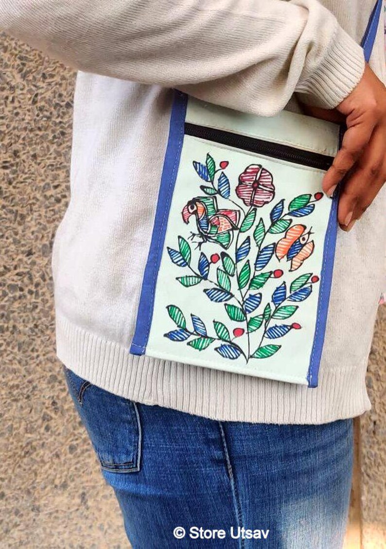 May include: A rectangular crossbody bag with a light green base, blue trim, and a black zipper closure. The front features a hand-drawn illustration of a bird, a flower, and a butterfly surrounded by green leaves and small red berries.