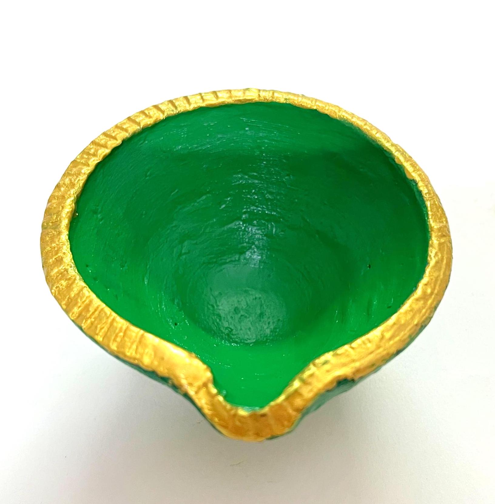 May include: A small, round, bowl-shaped diya lamp with a vibrant green interior and exterior. The rim of the lamp is painted with a textured, metallic gold finish, featuring a small pointed spout at the front edge for a wick to be placed.