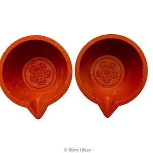 May include: Two terracotta oil lamps with a flower design in the center. The lamps are a warm orange-red color and have a small spout. The lamps are likely used for religious or decorative purposes.
