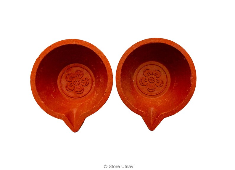 May include: Two terracotta oil lamps with a flower design in the center. The lamps are a warm orange-red color and have a small spout. The lamps are likely used for religious or decorative purposes.