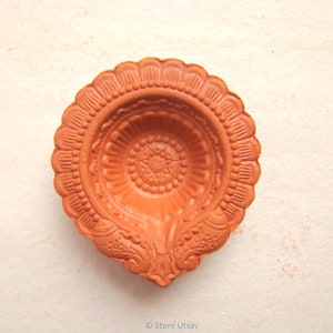 Pode incluir: Uma diya de terracota, ou l&acirc;mpada a &oacute;leo, com um design floral detalhado. A l&acirc;mpada tem uma cor castanho-alaranjada quente e tem uma borda ondulada. O design central apresenta um padr&atilde;o de estrela. A l&acirc;mpada &eacute; provavelmente usada para fins religiosos ou decorativos.