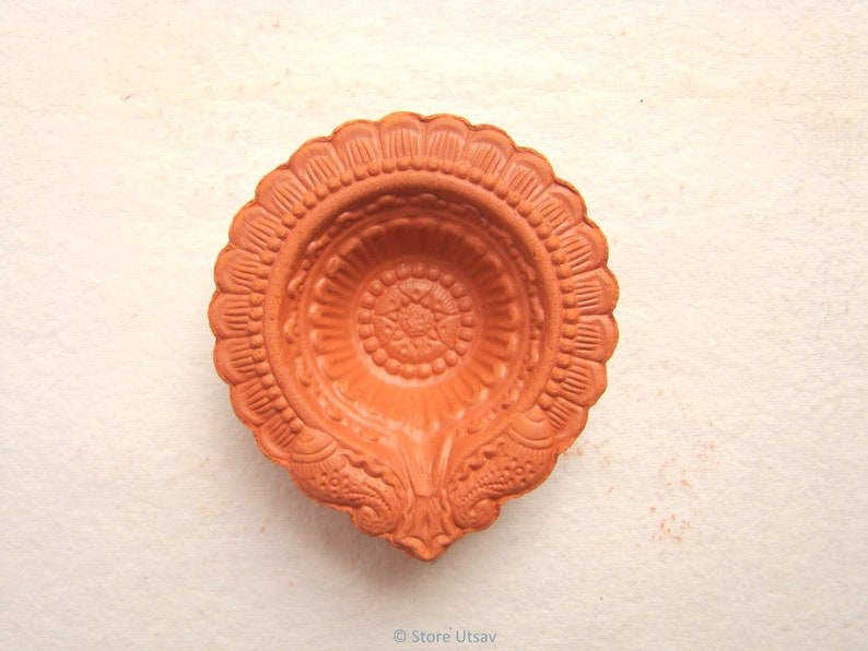 Pode incluir: Uma diya de terracota, ou l&acirc;mpada a &oacute;leo, com um design floral detalhado. A l&acirc;mpada tem uma cor castanho-alaranjada quente e tem uma borda ondulada. O design central apresenta um padr&atilde;o de estrela. A l&acirc;mpada &eacute; provavelmente usada para fins religiosos ou decorativos.