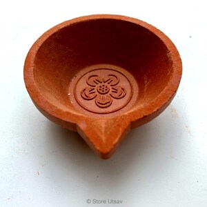 May include: A small, terracotta-colored oil lamp with a flower design in the center. The lamp has a rounded bowl shape with a pointed spout. The lamp is likely used for religious or decorative purposes.