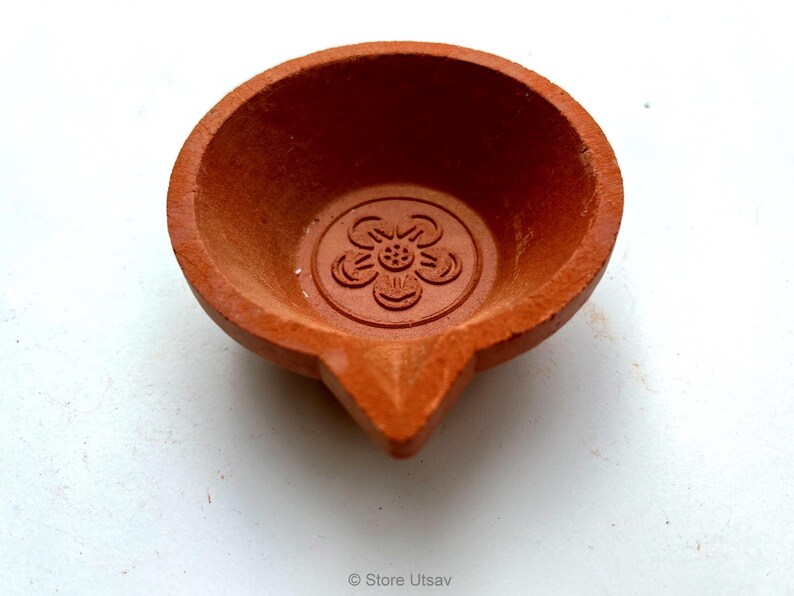 May include: A small, terracotta-colored oil lamp with a flower design in the center. The lamp has a rounded bowl shape with a pointed spout. The lamp is likely used for religious or decorative purposes.