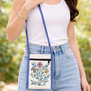 May include: A small, rectangular off-white canvas crossbody bag featuring a blue trim and a long blue shoulder strap. The front of the bag displays a colorful embroidered design of a flower, a bird, and a butterfly with green leaves.