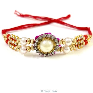 May include: An ornate bracelet with a large, central pearl surrounded by clear crystals. The bracelet features gold-colored accents, smaller pearls, and red and gold decorative elements. Red string forms the bracelet's ties.