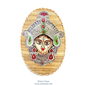 Puede incluir: Un tapiz ovalado para colgar en la pared pintado a mano que presenta una representación colorida de la diosa hindú Durga. La obra de arte está pintada en una estera de bambú tejida con un borde de cuerda blanca.