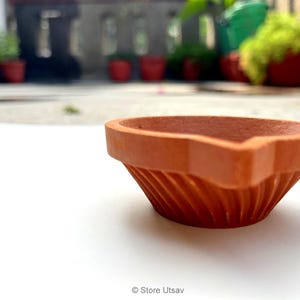 May include: A terracotta diya, a traditional oil lamp, sits on a white surface. The lamp has a wide, fluted bowl and a pointed spout for the wick. The background is blurred, with potted plants visible.