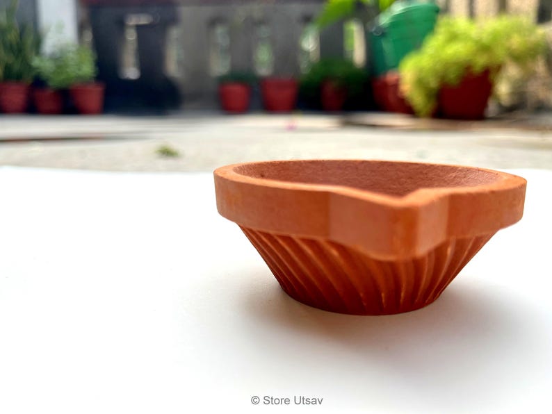 May include: A terracotta diya, a traditional oil lamp, sits on a white surface. The lamp has a wide, fluted bowl and a pointed spout for the wick. The background is blurred, with potted plants visible.