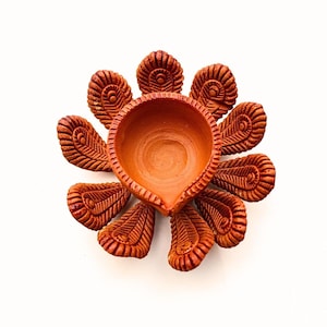 May include: A terracotta oil lamp featuring a central circular bowl surrounded by ten decorative petals arranged in a radial pattern. Each petal displays a textured, embossed feather-like design with fine lines and a small circular detail at the base.