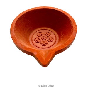May include: A terracotta-colored diya, a traditional oil lamp, with a flower design in the center. The lamp has a rounded bowl shape with a pointed spout. The diya is used for religious and festive occasions.