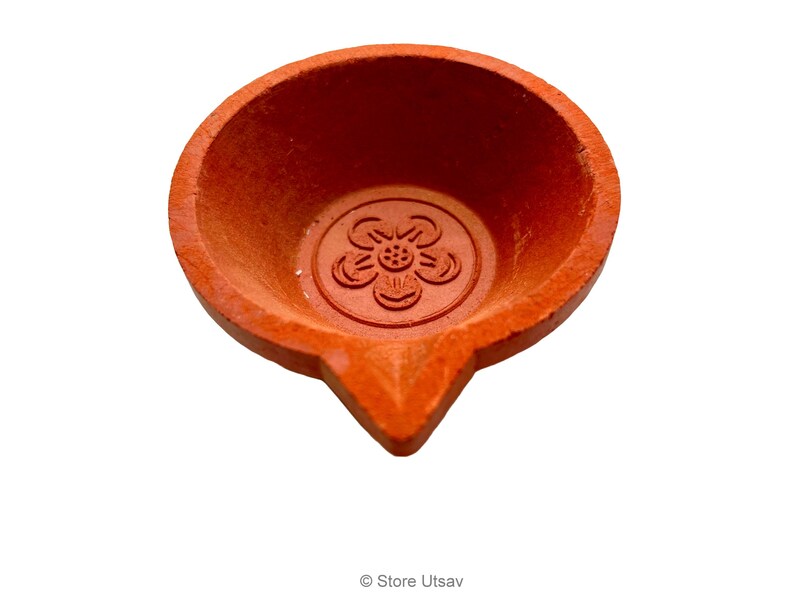 May include: A terracotta-colored diya, a traditional oil lamp, with a flower design in the center. The lamp has a rounded bowl shape with a pointed spout. The diya is used for religious and festive occasions.