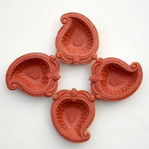 May include: Four terracotta diya lamps, each with a decorative design, arranged in a cross pattern on a white background.