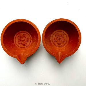 May include: Two terracotta oil lamps, each with a flower design in the center. The lamps are a warm orange-red color and have a small spout for the wick. The lamps are likely used for religious or decorative purposes.