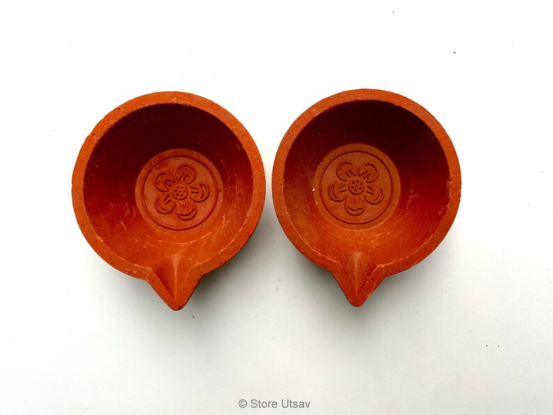 May include: Two terracotta oil lamps, each with a flower design in the center. The lamps are a warm orange-red color and have a small spout for the wick. The lamps are likely used for religious or decorative purposes.