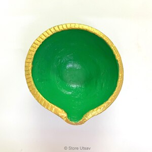 May include: A round, bowl-shaped diya lamp featuring a vibrant green interior and a textured gold-painted rim. The vessel has a small, distinct spout at the front edge for a wick, with a smooth, matte finish on the inner surface and a metallic gold border.