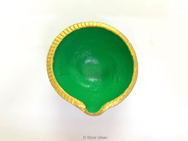 May include: A round, bowl-shaped diya lamp featuring a vibrant green interior and a textured gold-painted rim. The vessel has a small, distinct spout at the front edge for a wick, with a smooth, matte finish on the inner surface and a metallic gold border.