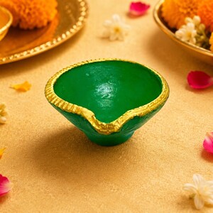 May include: A small, bowl-shaped oil lamp, known as a diya, featuring a vibrant green interior and exterior. The rim of the lamp is decorated with a textured, metallic gold painted edge, providing a distinct contrast to the smooth green finish.