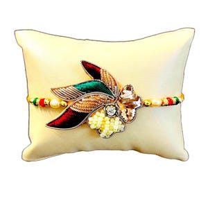 Puede incluir: Una pulsera decorativa con un diseño floral, con detalles en rojo, verde y dorado. La pulsera está adornada con un cristal grande y transparente y pequeñas cuentas blancas. Descansa sobre una almohada color crema.