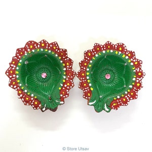 May include: Two green and red painted diya lamps with white and yellow accents. The diyas are decorated with a floral design and have a small pink jewel in the center.