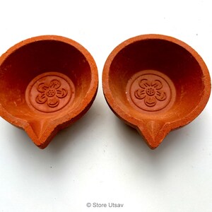 May include: Two terracotta-colored oil lamps, each with a flower design embossed in the center. The lamps have a rounded bowl shape with a pointed spout. The lamps are on a white background.