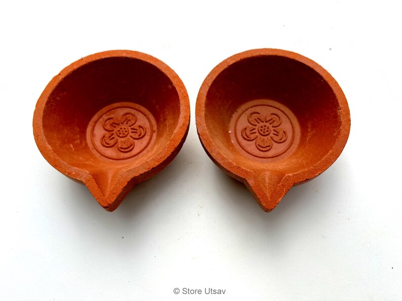 May include: Two terracotta-colored oil lamps, each with a flower design embossed in the center. The lamps have a rounded bowl shape with a pointed spout. The lamps are on a white background.