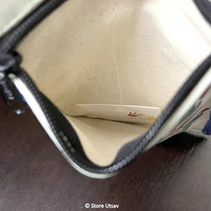 May include: An open rectangular pouch with a cream-colored interior lining and a black zipper closure. The exterior features blue fabric edges and a white surface with a partial graphic print. A small white card with a hand print logo is visible inside.