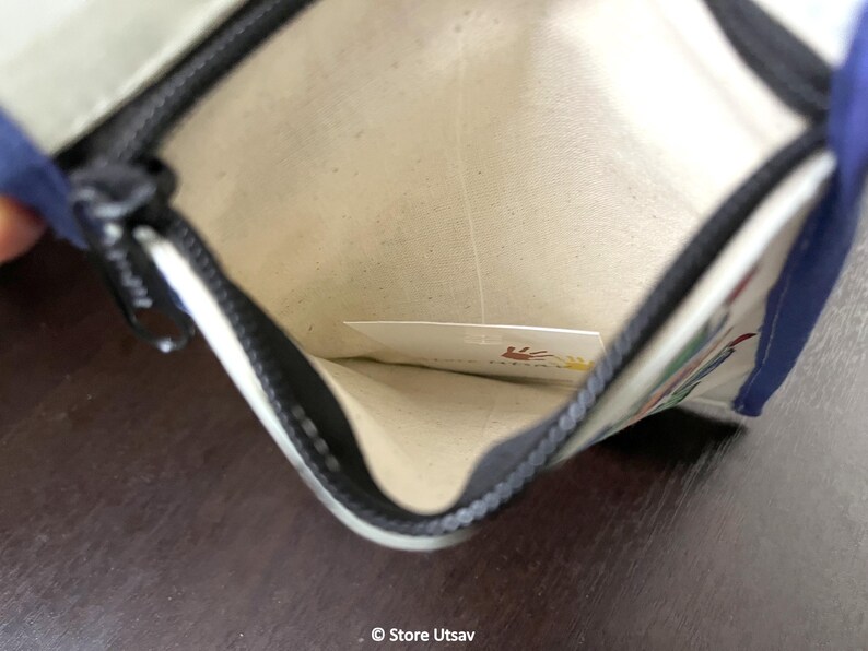 May include: An open rectangular pouch with a cream-colored interior lining and a black zipper closure. The exterior features blue fabric edges and a white surface with a partial graphic print. A small white card with a hand print logo is visible inside.