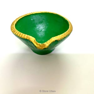 May include: A small, bowl-shaped diya lamp painted in a vibrant green color. The circular rim features a textured, scalloped edge that is highlighted with a metallic gold paint. A small spout for a wick is located at the front of the lamp.