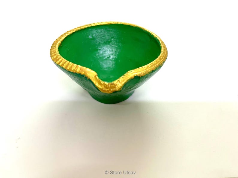 May include: A small, bowl-shaped diya lamp painted in a vibrant green color. The circular rim features a textured, scalloped edge that is highlighted with a metallic gold paint. A small spout for a wick is located at the front of the lamp.