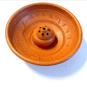 May include: A round, terracotta-coloured incense burner bowl featuring a central raised dome with multiple small circular perforations. The interior surface is decorated with a series of etched, angled dash marks and concentric circular lines.
