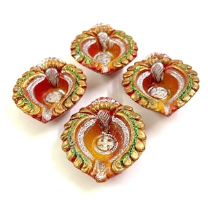 May include: Four decorative, heart-shaped oil lamps. The lamps are painted with red, orange, gold, silver, and green accents. The lamps are arranged in a square formation against a white background. The lamps are likely used for religious or cultural celebrations.